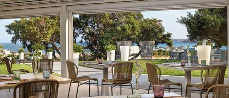 Un área de comedor al aire libre con sillas y mesas de mimbre, con vistas a un paisaje marítimo y vegetación.