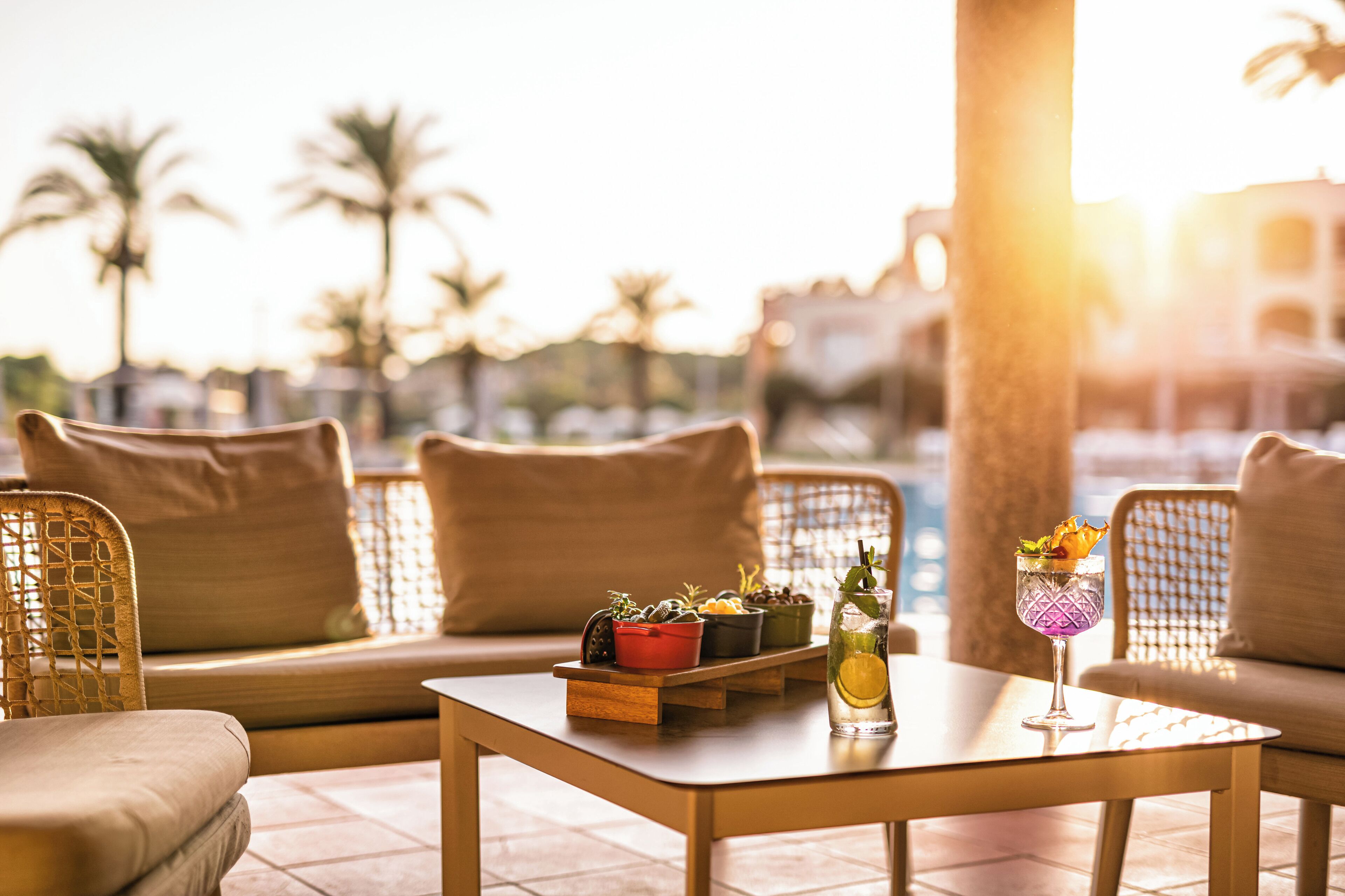 A sunlit outdoor seating area with wicker furniture, a wooden table with a drink and potted plants, and a view of palm trees and buildings in the background.