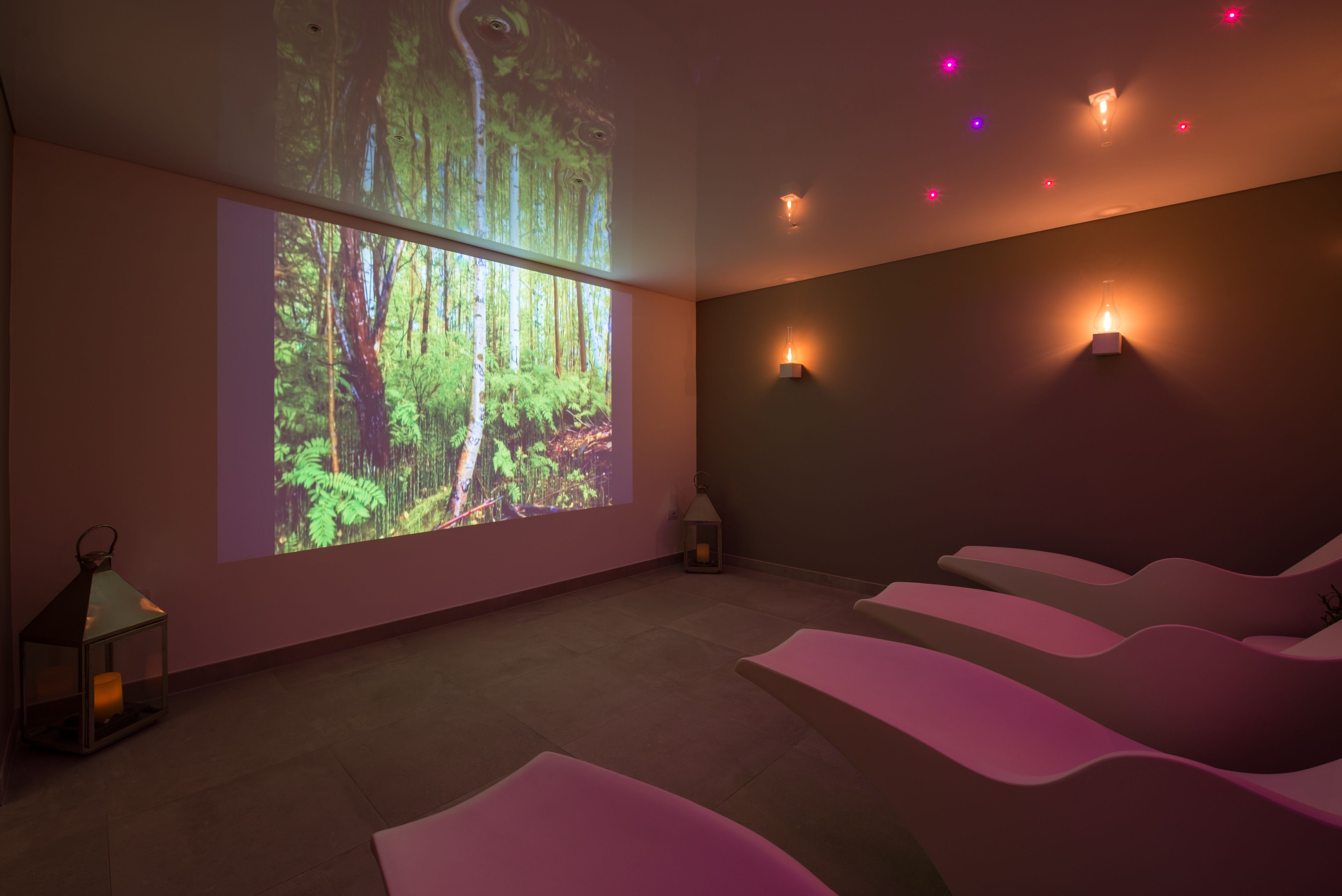 A room with a large projection screen displaying a forest scene, cushioned seating, wall-mounted lights, and a lantern.