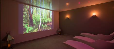 Habitación con una pantalla grande que muestra un paisaje forestal, asientos acolchados, luces empotradas en la pared y un farol.