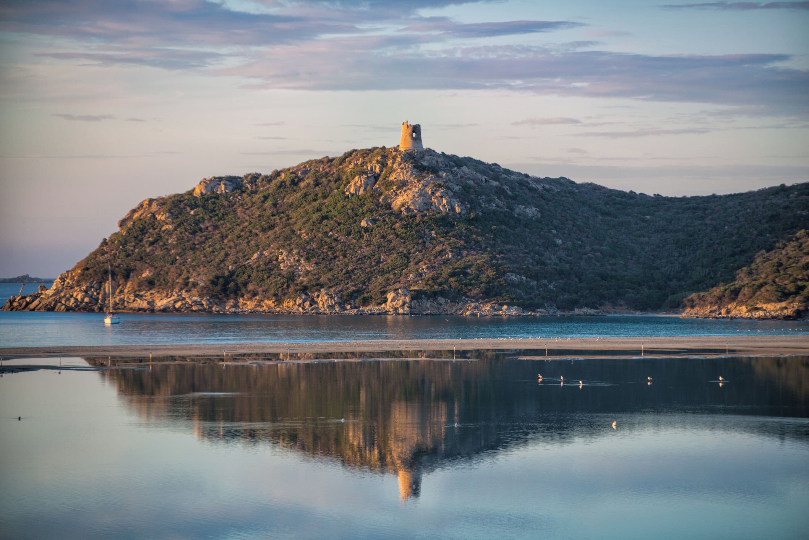 Kustlandschap met een heuvel waarop een toren staat, kalm water dat het tafereel weerspiegelt, en een heldere lucht.
