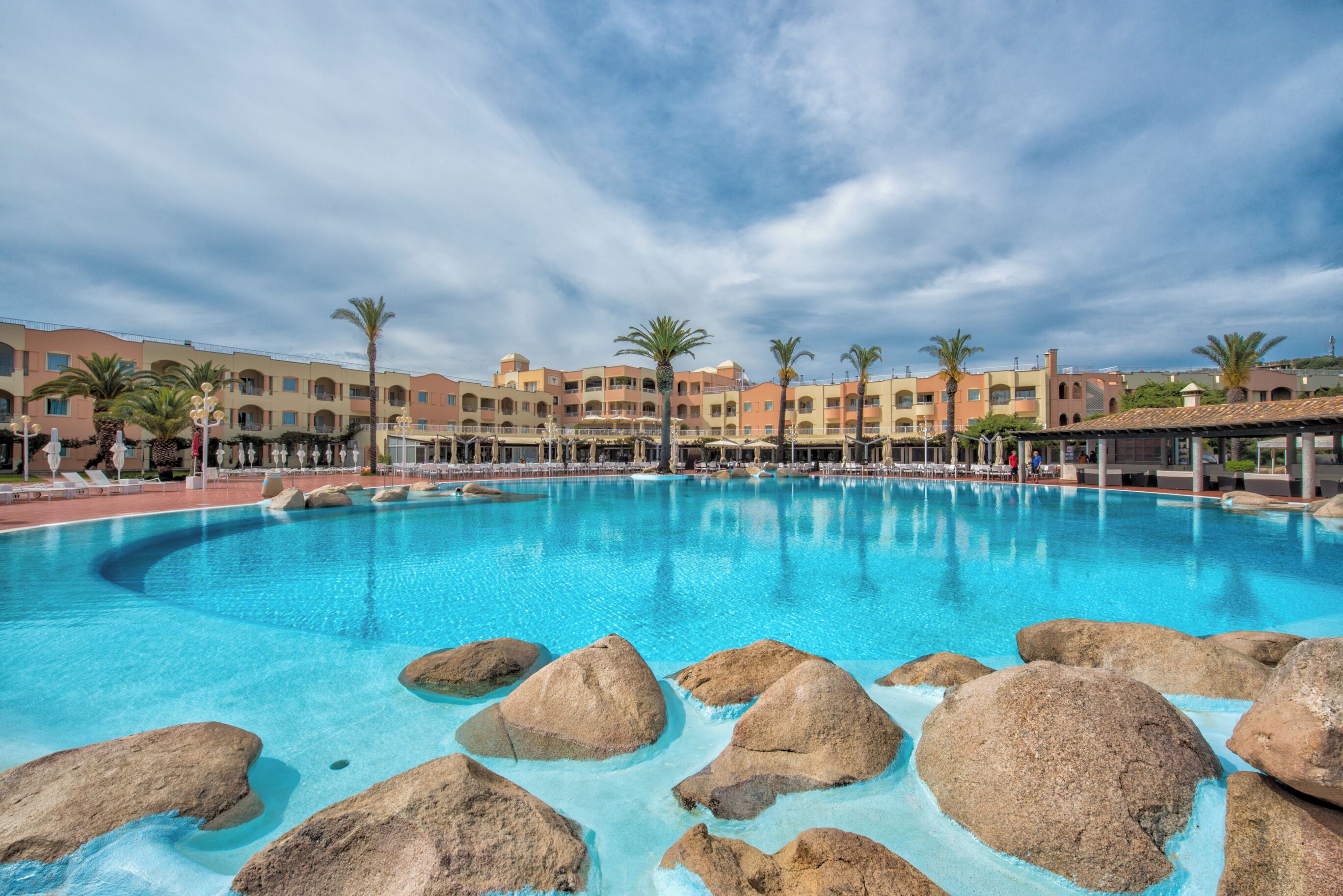 A large outdoor swimming pool with rocks, surrounded by palm trees and hotel buildings.