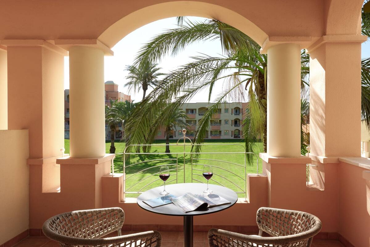 A balcony with two wicker chairs, a table with two wine glasses and a menu, and a view of a building and palm trees.