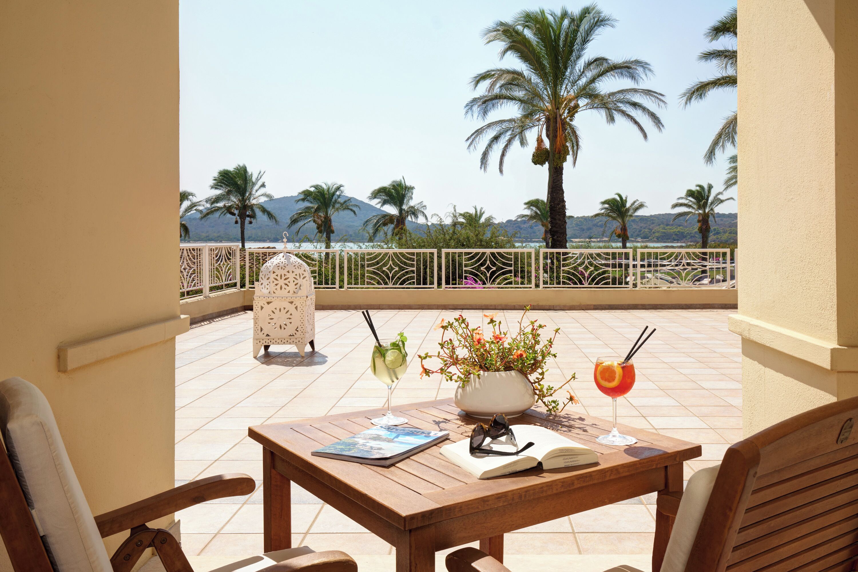 A wooden table with a book, a pair of glasses, and two drinks on a balcony overlooking palm trees and mountains.