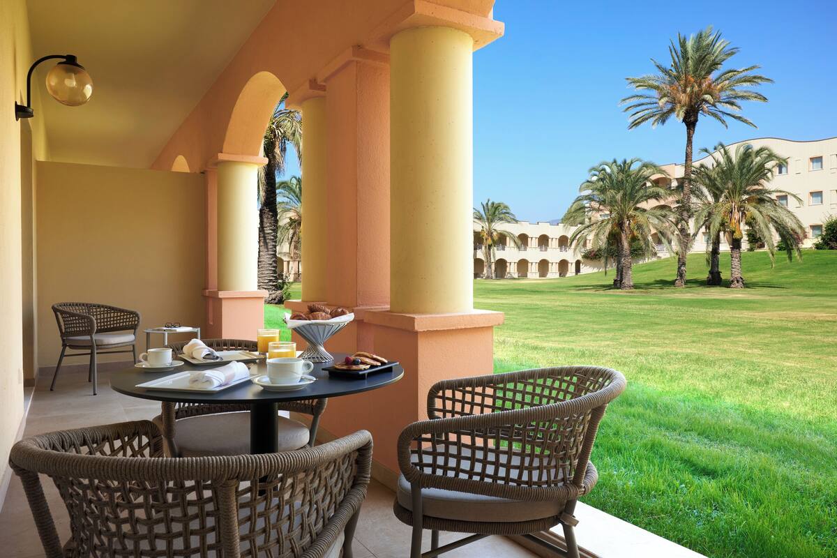 A patio with wicker chairs, a table set with breakfast items, and a view of a well-maintained lawn and palm trees.