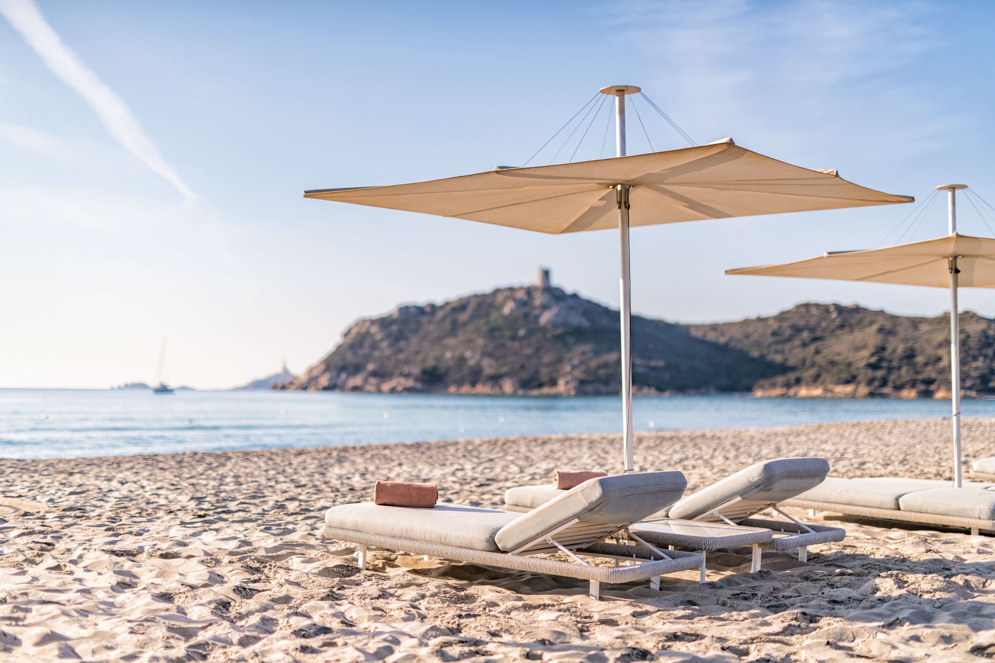 A beach with sun loungers and umbrellas, a sandy shore, and a distant hill.
