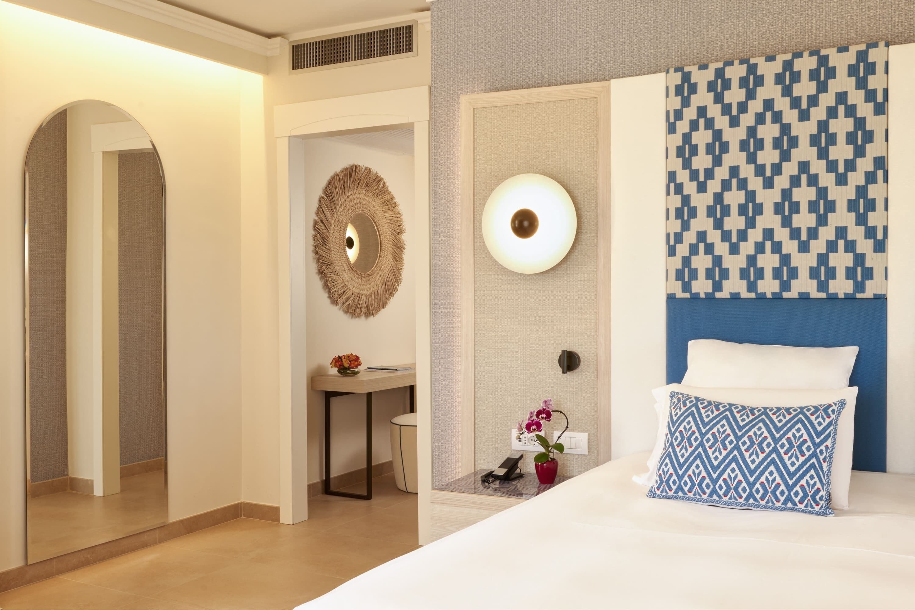A hotel room with a bed, a desk, a mirror, and a wall-mounted light fixture.