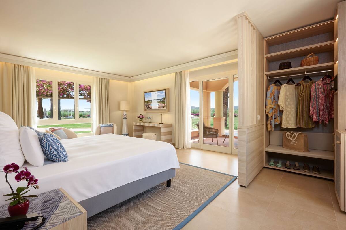 A hotel room with a large bed, a desk, a chair, and a wardrobe. There is a view of the outdoors through the windows.