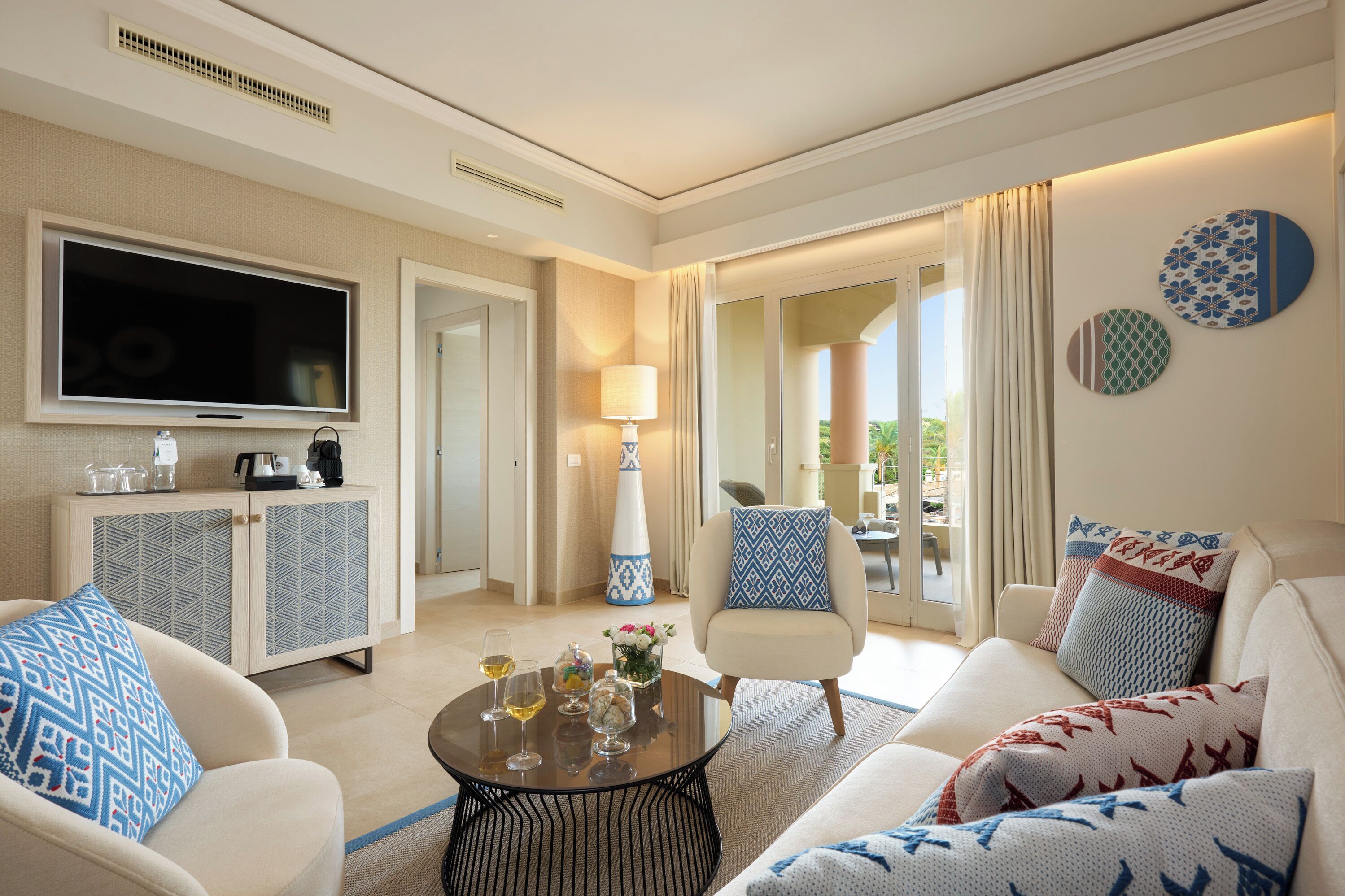 A modern hotel room with a flat-screen TV, a sofa, a coffee table with drinks, a cabinet, and a view of the outdoors.