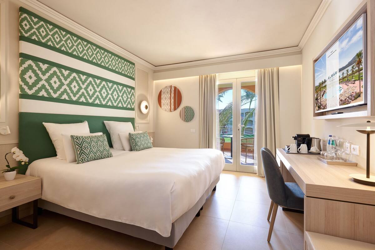A hotel room with a bed, a desk, a chair, a TV, and a balcony with curtains.