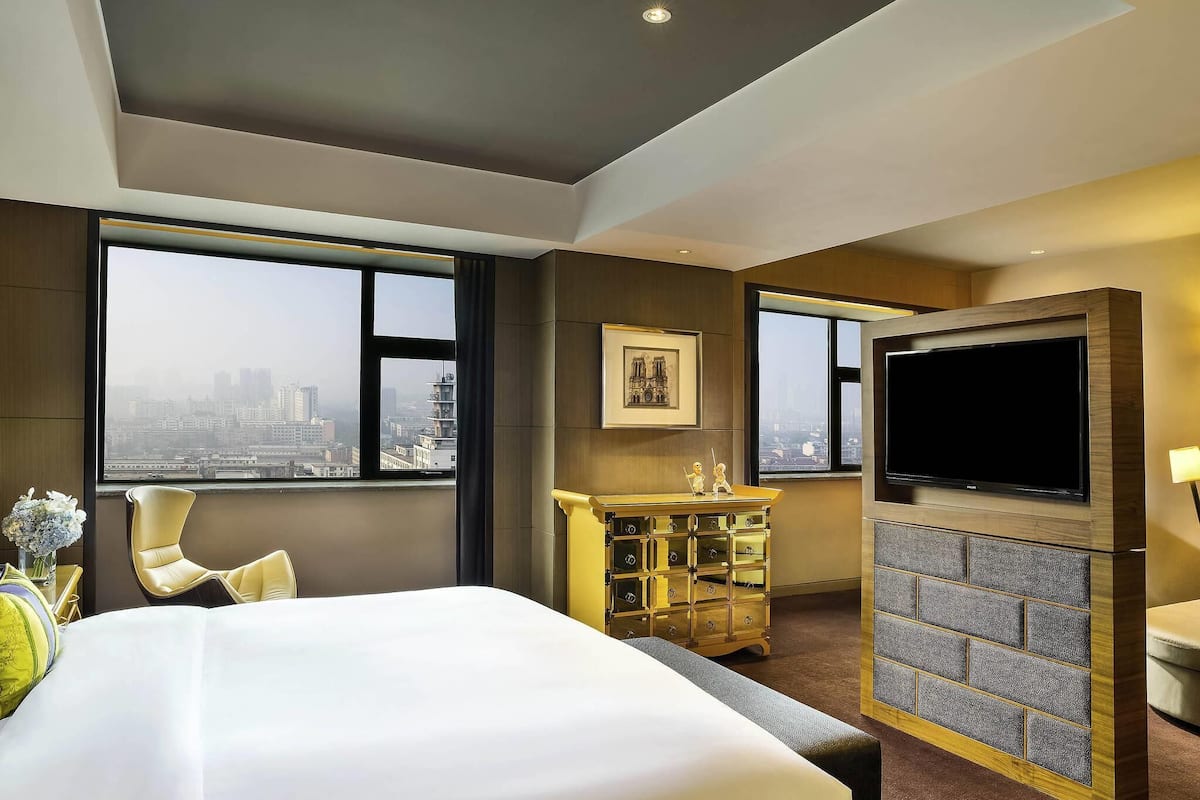 Habitación de hotel con cama, televisión, chimenea, escritorio y ventanas amplias con vistas a la ciudad.
