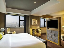 Habitación de hotel con cama, televisión, chimenea, escritorio y ventanas amplias con vistas a la ciudad.