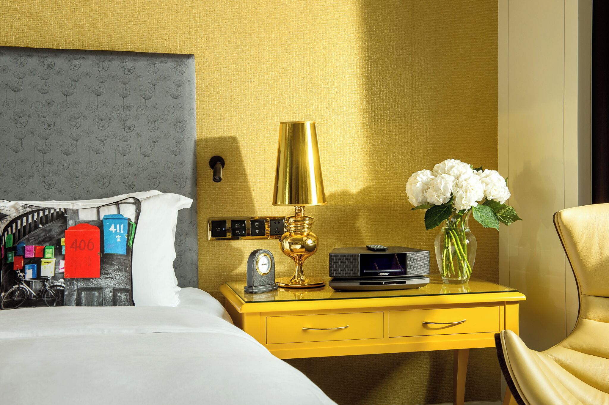 A hotel room with a bed, a yellow nightstand with a lamp, a vase with flowers, and a chair.