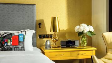 A hotel room with a bed, a yellow nightstand with a lamp, a vase with flowers, and a chair.