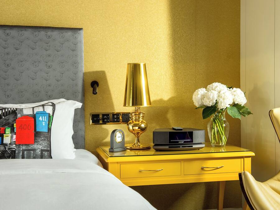A hotel room with a bed, a yellow nightstand with a lamp, a vase with flowers, and a chair.