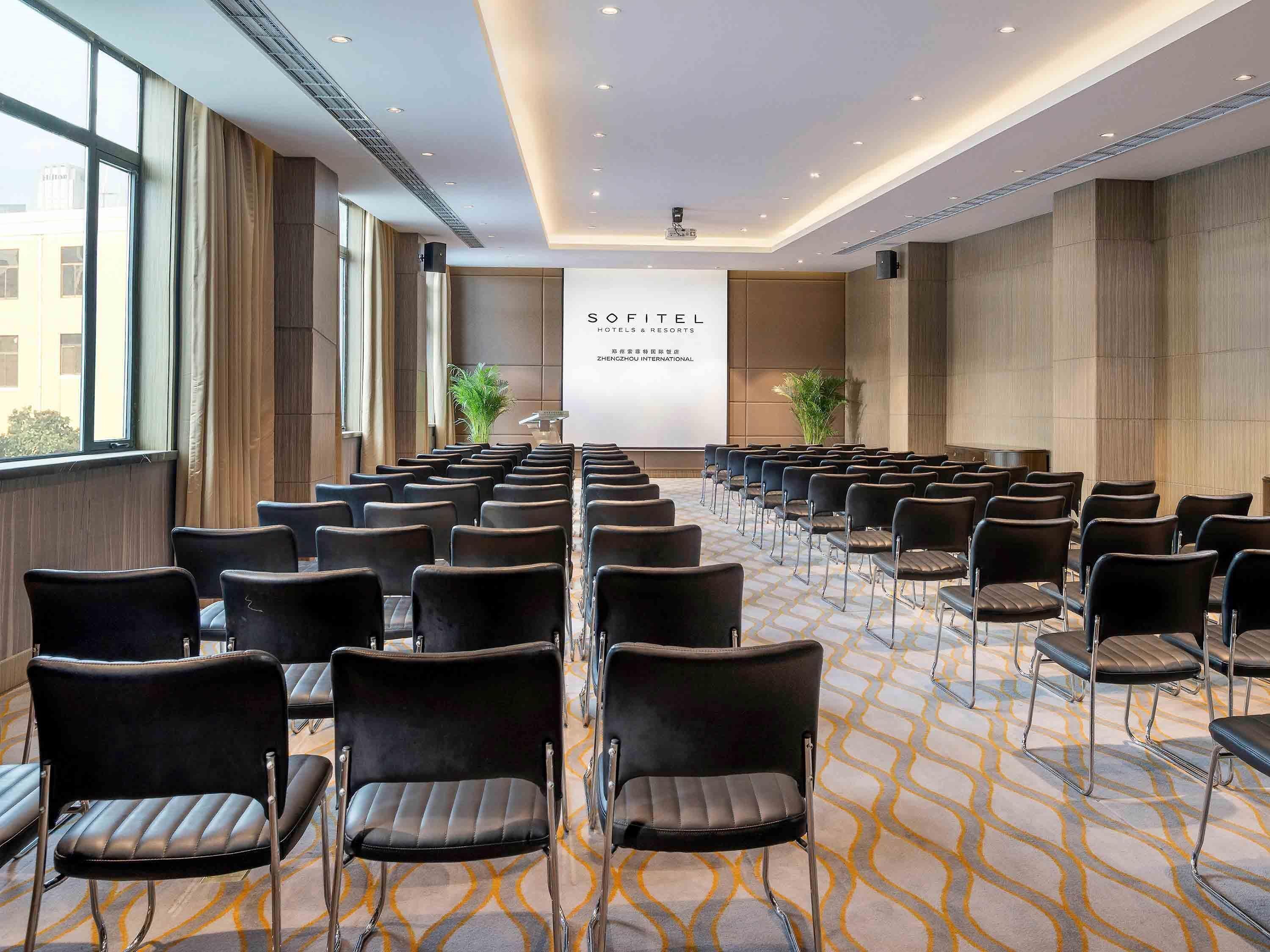 Une salle de conférence avec des rangées de chaises, un pupitre et un écran de projection affichant des informations sur l’hôtel.