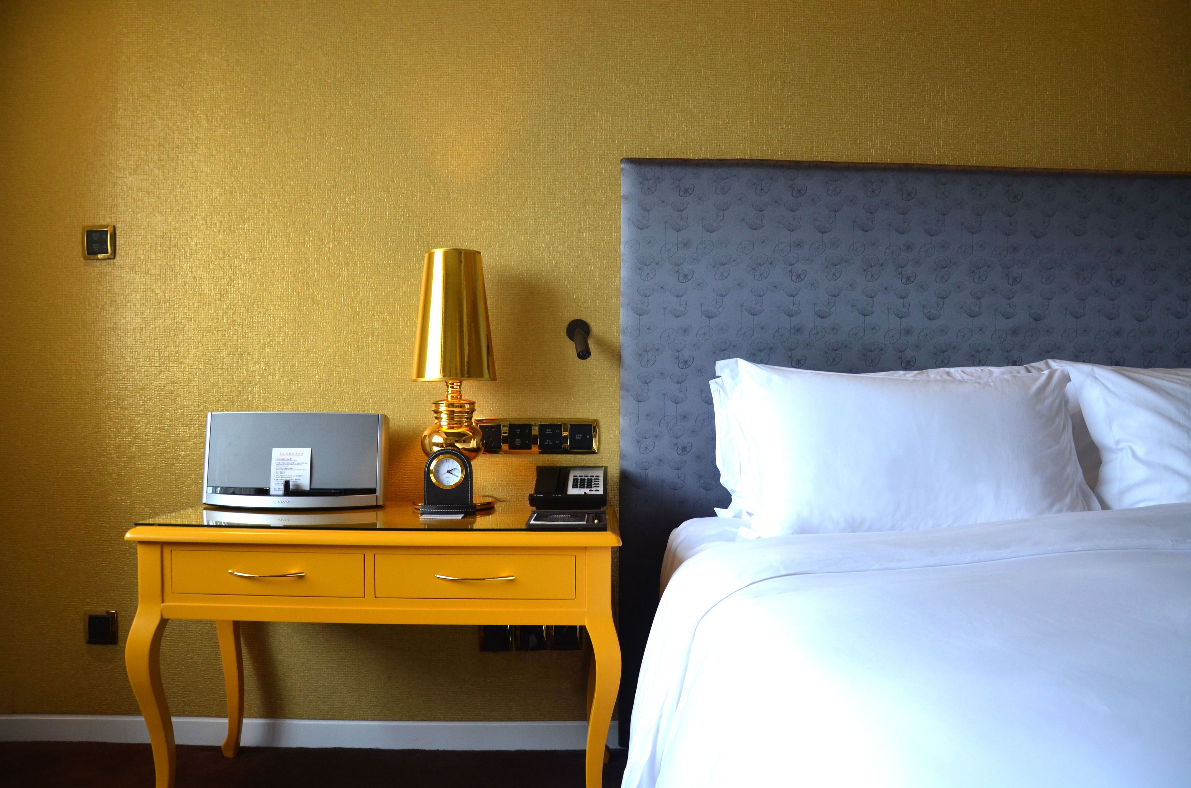 A hotel room with a bed, a nightstand with a lamp, a clock, and a small safe.