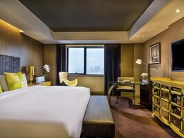 Ein Hotelzimmer mit einem großen Bett, einem Schreibtisch, einem Sessel, einer Kommode und einem Fenster mit Blick auf die Stadt.