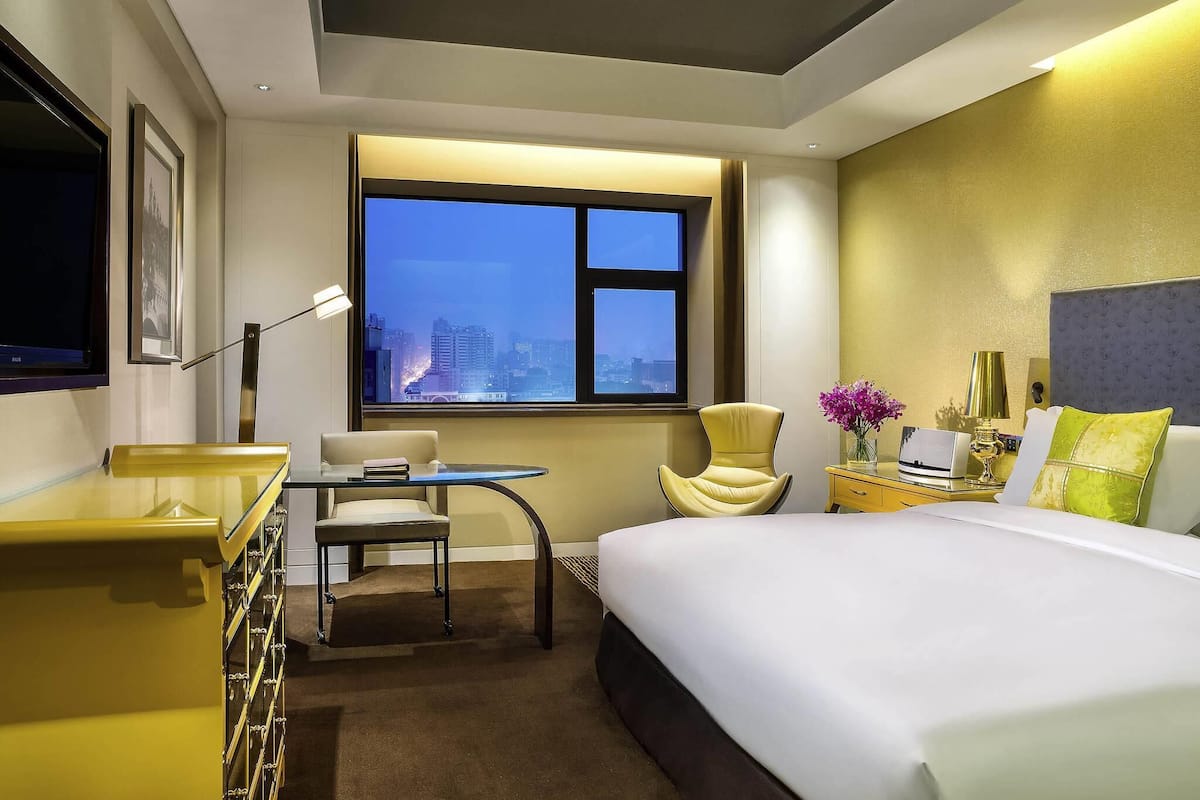 Una habitación de hotel con cama, escritorio, silla, ventana con vista a la ciudad y mesita de noche con lámpara.