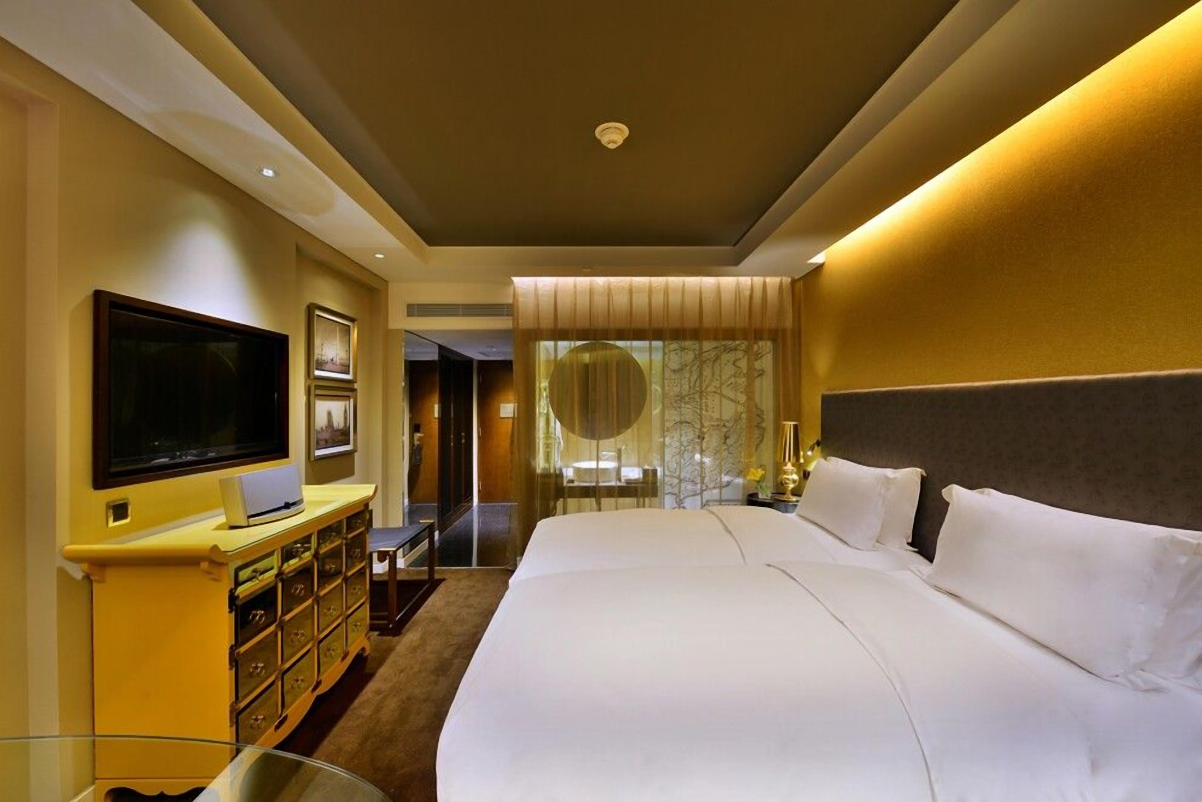 A hotel room with a large bed, a flat-screen TV, a minibar, and a view of a cityscape through the window.
