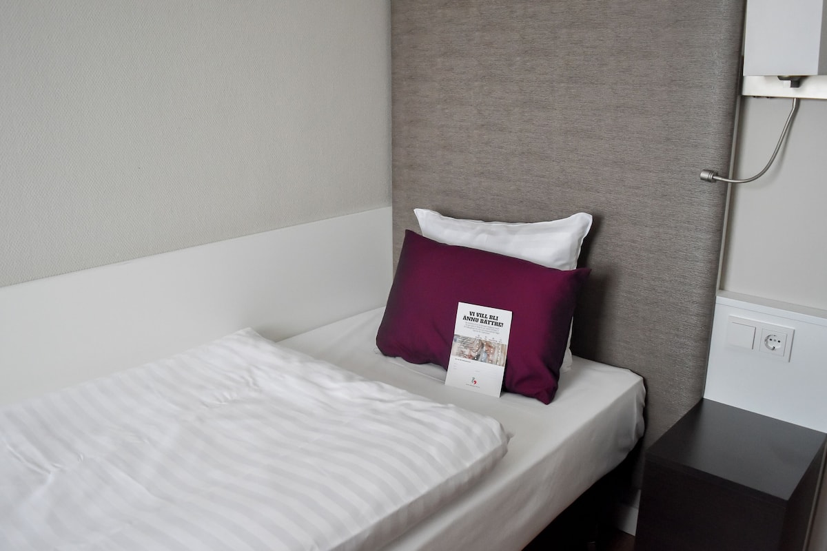 A neatly made bed with white and striped bedding, a dark headboard, and a small bedside table with a power outlet.