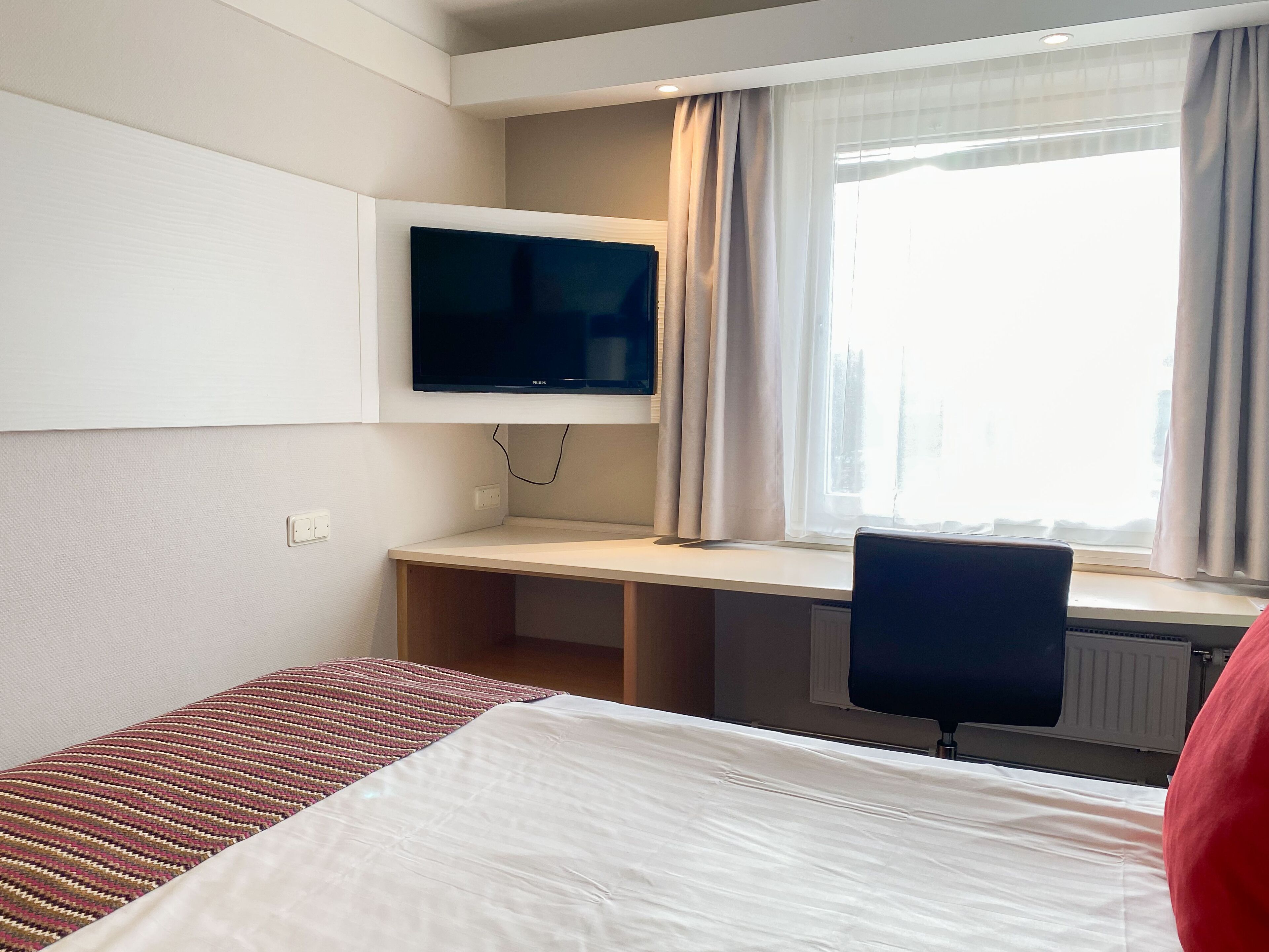 Ett hotellrum med en säng, ett skrivbord, en stol, en tv och ett fönster med gardiner.