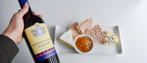 Una mano sosteniendo una botella de vino Montepulciano d'Abruzzo, un plato blanco con queso, galletas y un cuenco pequeño de mermelada.