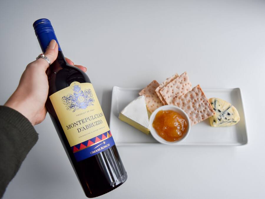 Una mano sosteniendo una botella de vino Montepulciano d'Abruzzo, un plato blanco con queso, galletas y un cuenco pequeño de mermelada.