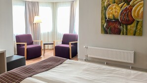Un cuarto de hotel con una cama, dos sillones morados, una mesita, una lámpara, un radiador y un cuadro de frutas en la pared.