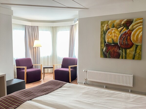 A hotel room with a bed, two purple armchairs, a small table, a lamp, a radiator, and a painting of fruits on the wall.