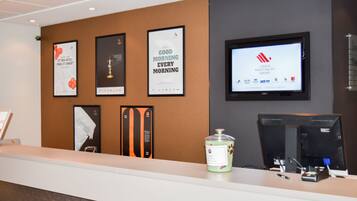 A hotel reception area with a counter, framed posters, a television screen, and a trash can.