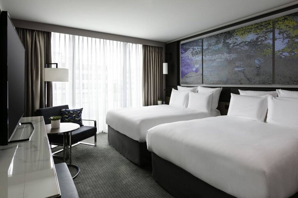A hotel room with two beds, a desk, a chair, a TV, and a large window with curtains.