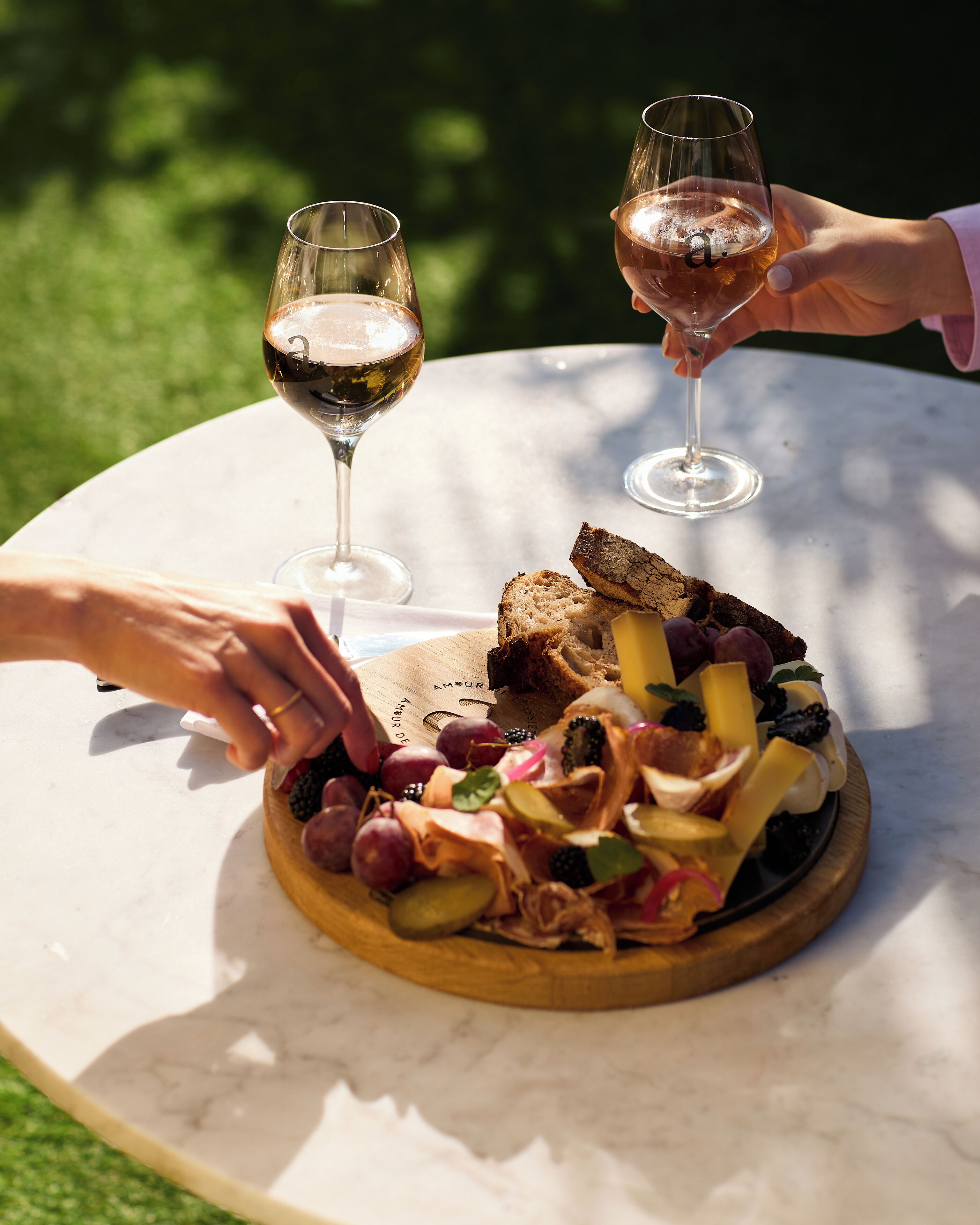 Une table ronde surmontée d’une planche en bois servant de présentoir pour de la charcuterie, avec deux verres à vin et une personne qui tend la main pour prendre un morceau de nourriture.
