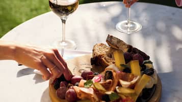 A round table with a wooden board holding a charcuterie board, two wine glasses, and a person reaching for a piece of food.