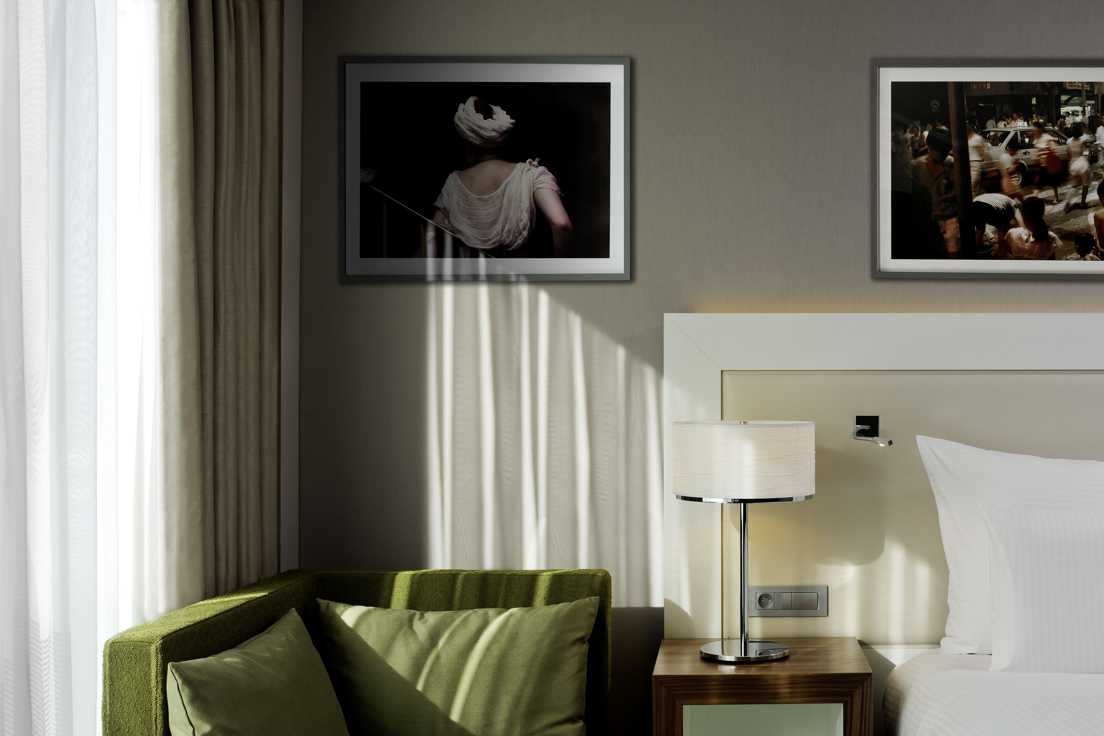 A hotel room with a bed, a nightstand with a lamp, a green armchair, and two framed pictures on the wall.