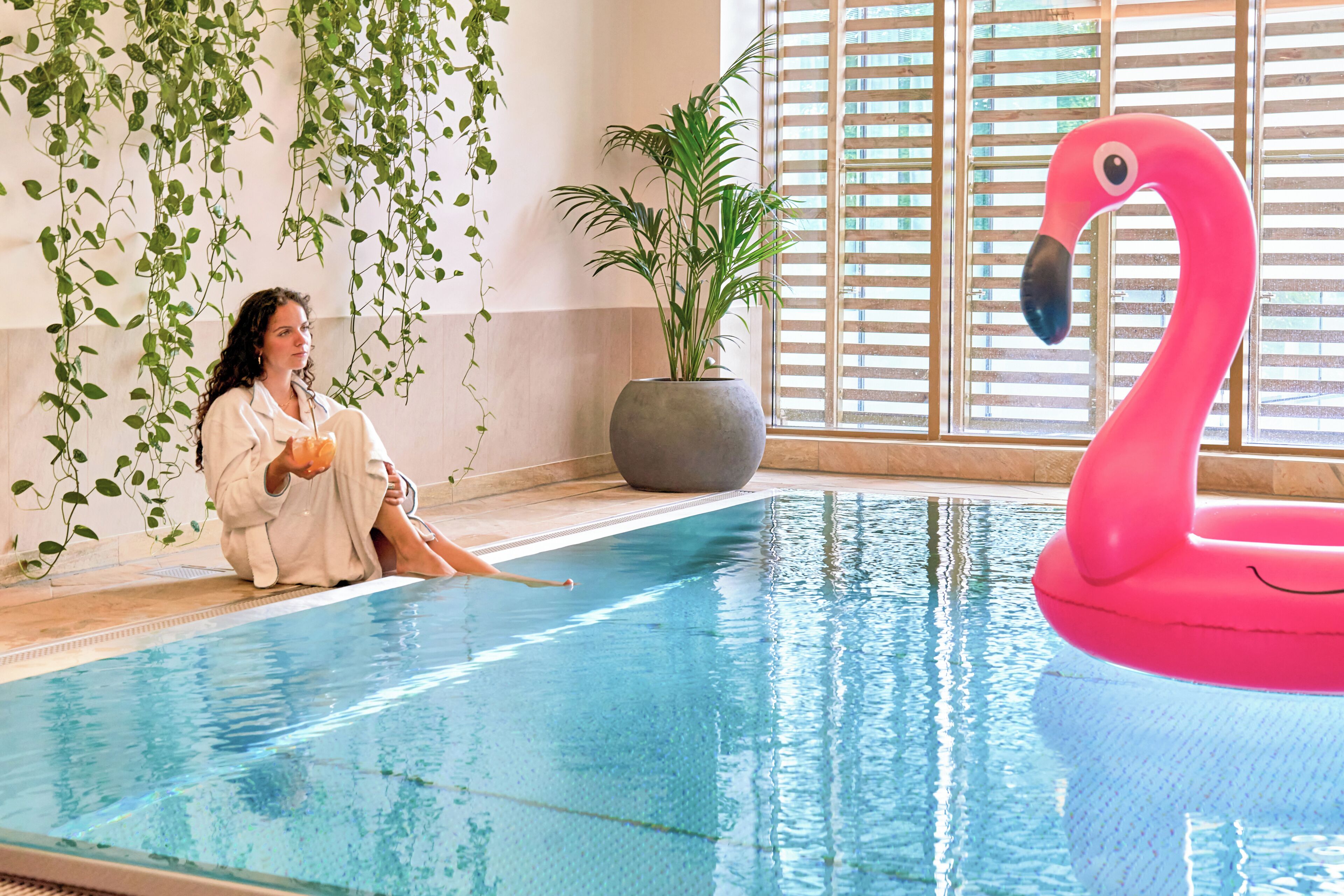 A person sitting by a pool with a flamingo float, indoor plants, and a potted plant.