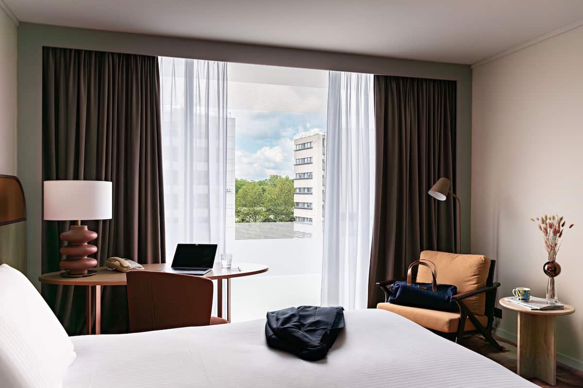 A hotel room with a bed, a desk with a laptop, a chair, a lamp, and a view of buildings and trees through the window.