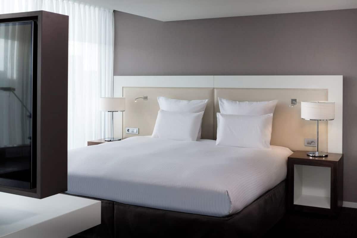 A modern hotel room with a large bed, two bedside tables with lamps, and a view of the city through a large window.