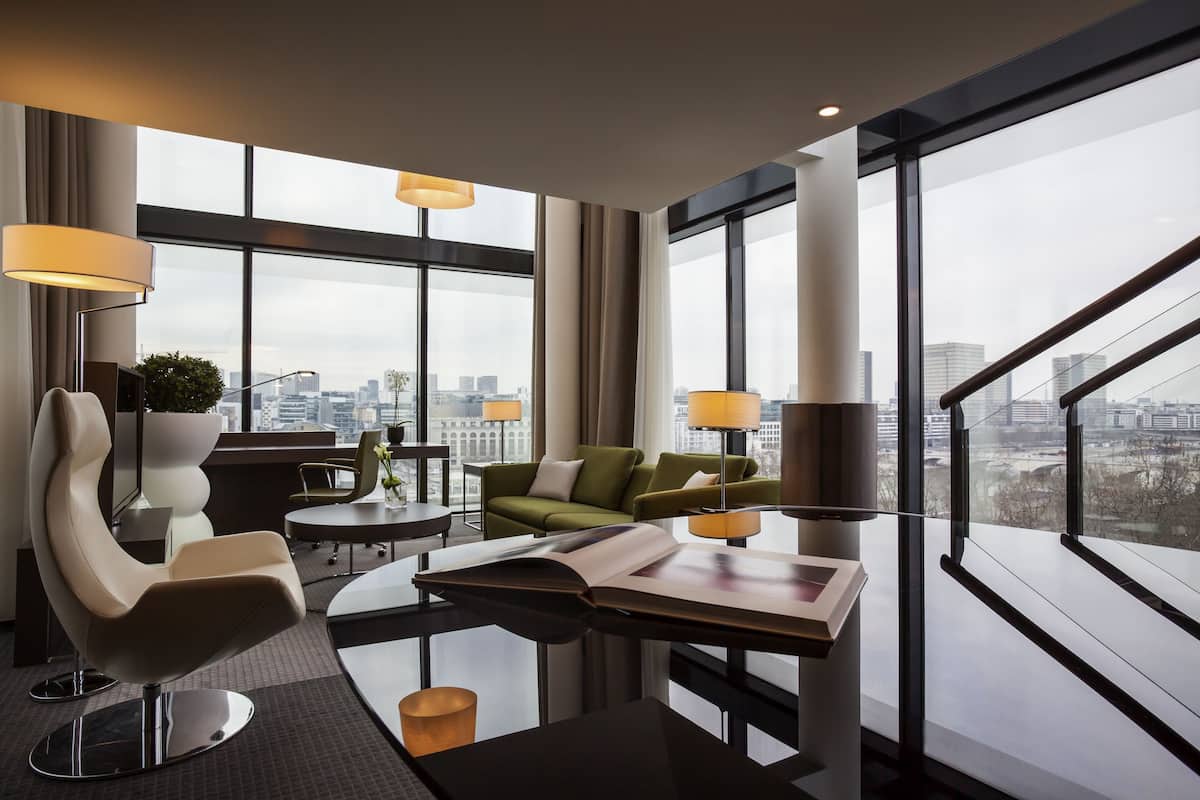 A modern hotel room with a large window, a green sofa, a white chair, and a glass coffee table.