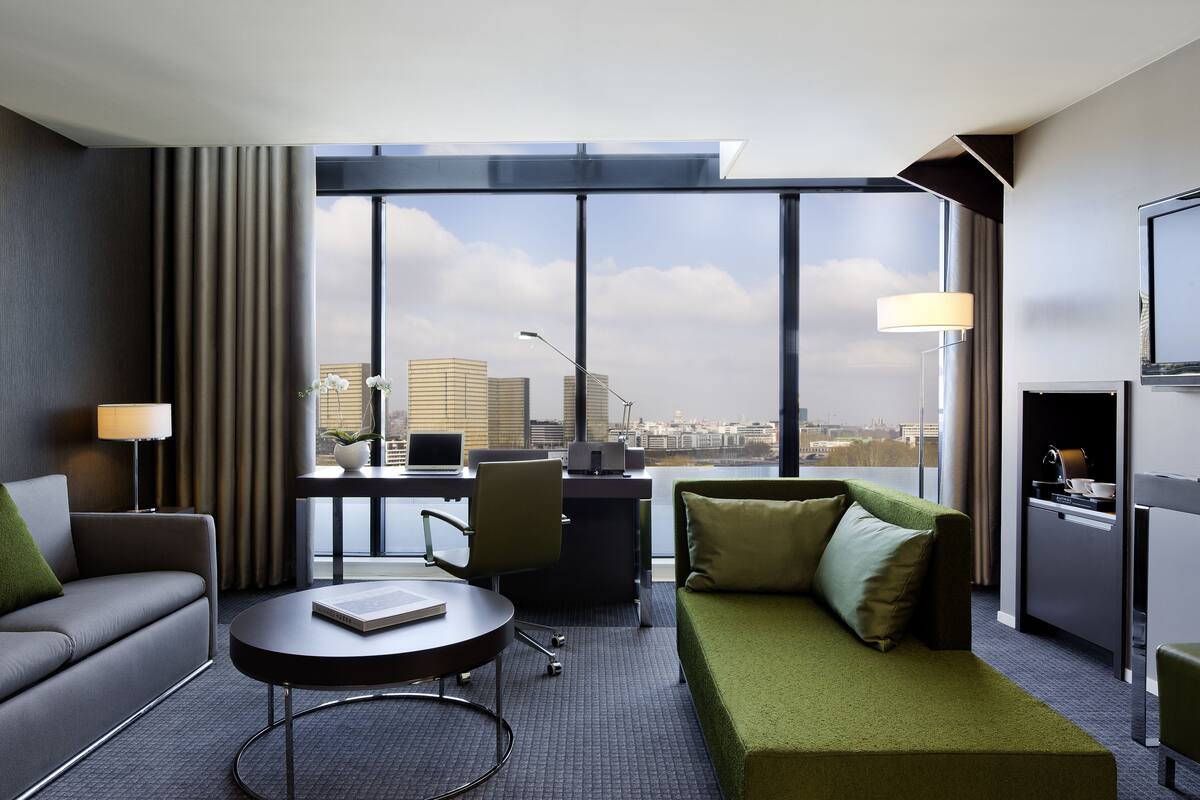 A modern hotel room with a sofa, a small table, a desk with a computer, and a view of the cityscape.