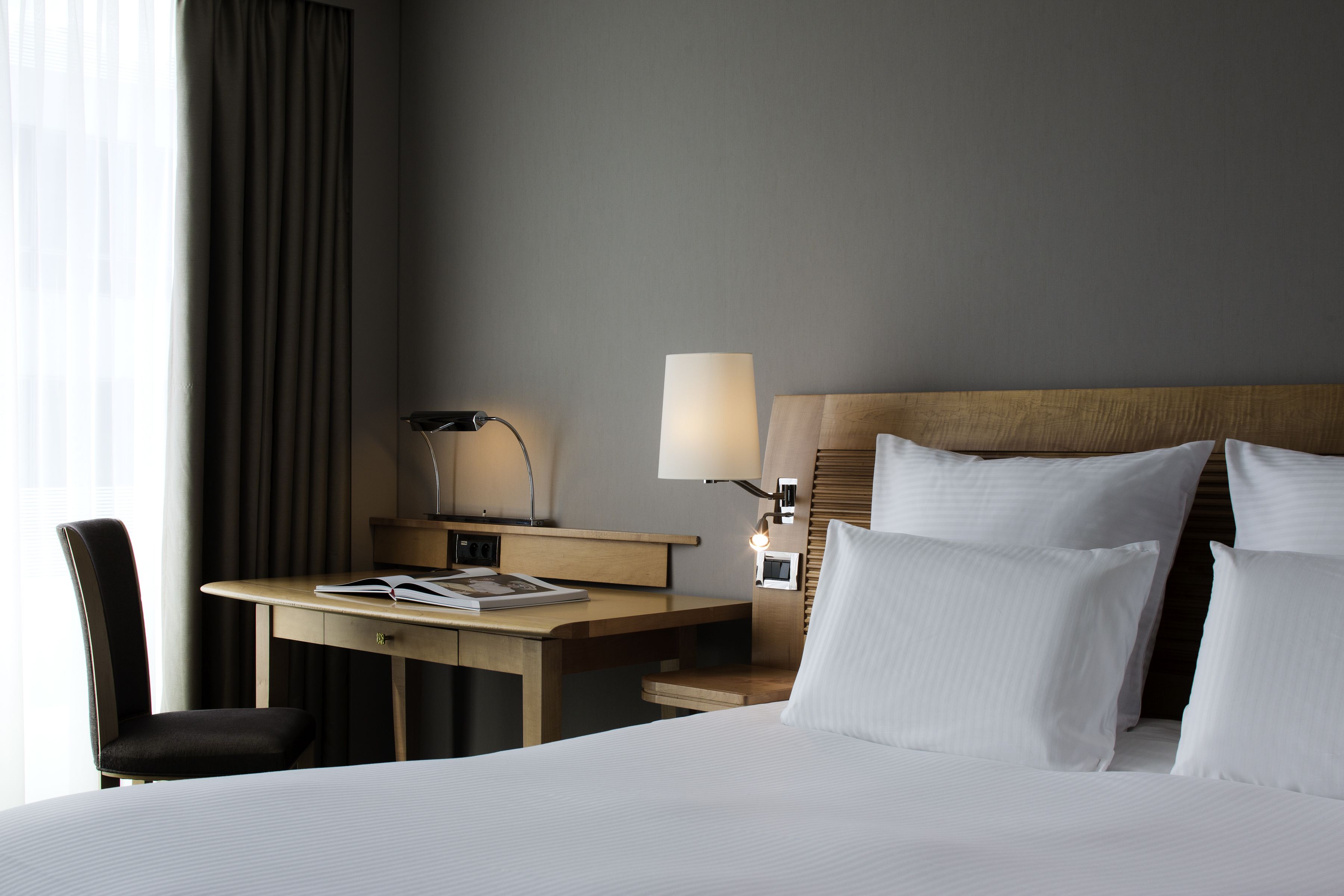 A hotel room with a bed, a desk with a lamp, a chair, and a nightstand with a lamp.