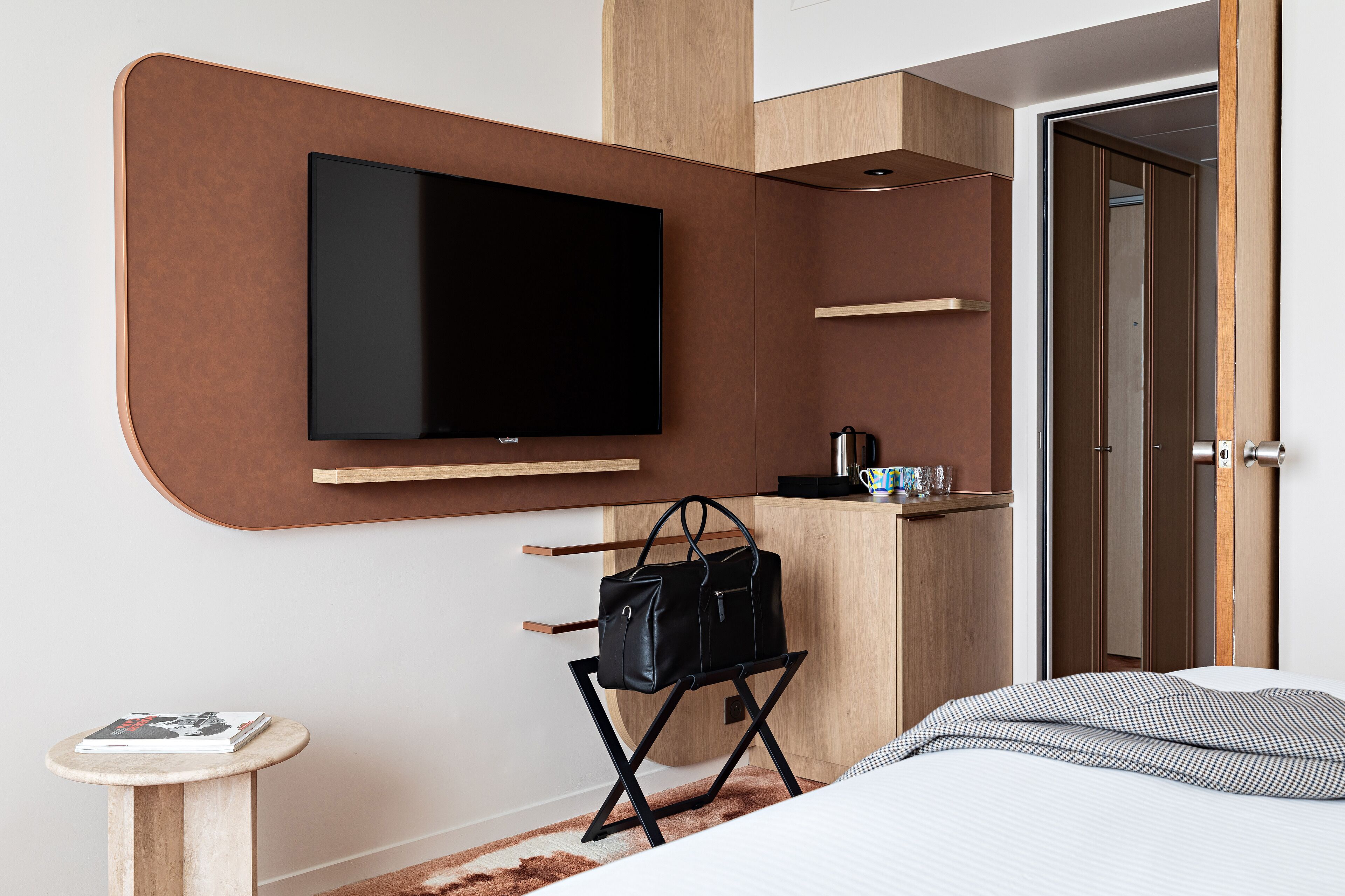 Una habitación de hotel moderna con un televisor de pantalla plana montado en un panel de madera, una mesita con una revista, un bolso de mano negro sobre un soporte y una cama con edredón estampado.