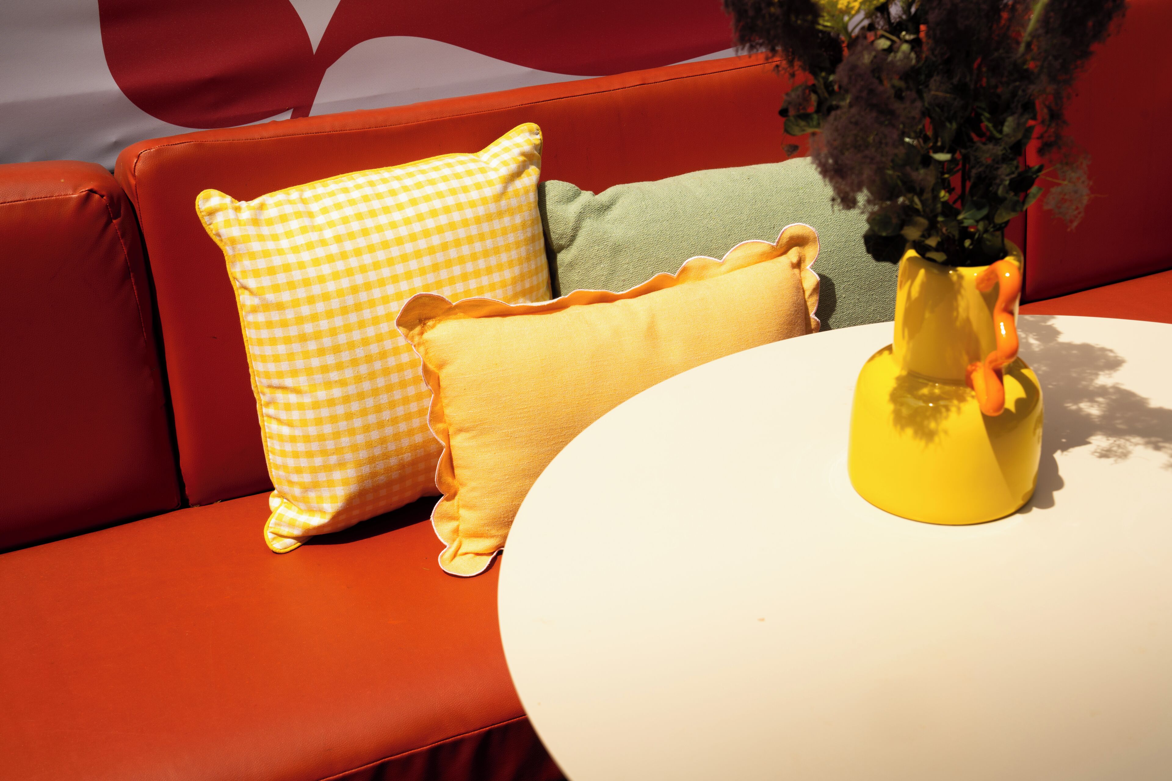 Un canapé rouge avec des coussins jaunes et verts, une table blanche et un vase jaune contenant une plante.