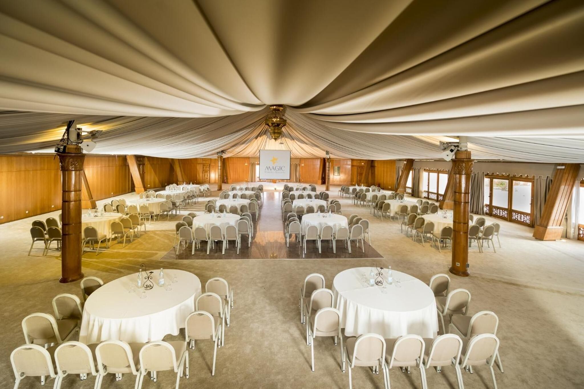 A large banquet hall with round tables and chairs, a stage at the front, and wooden paneling on the walls.