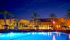 A swimming pool with lounge chairs and umbrellas, surrounded by palm trees and a building with large windows and warm lighting.