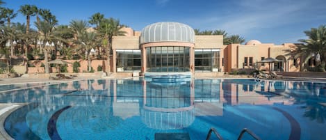 A swimming pool with a glass dome structure, surrounded by palm trees and a building with a beige facade.