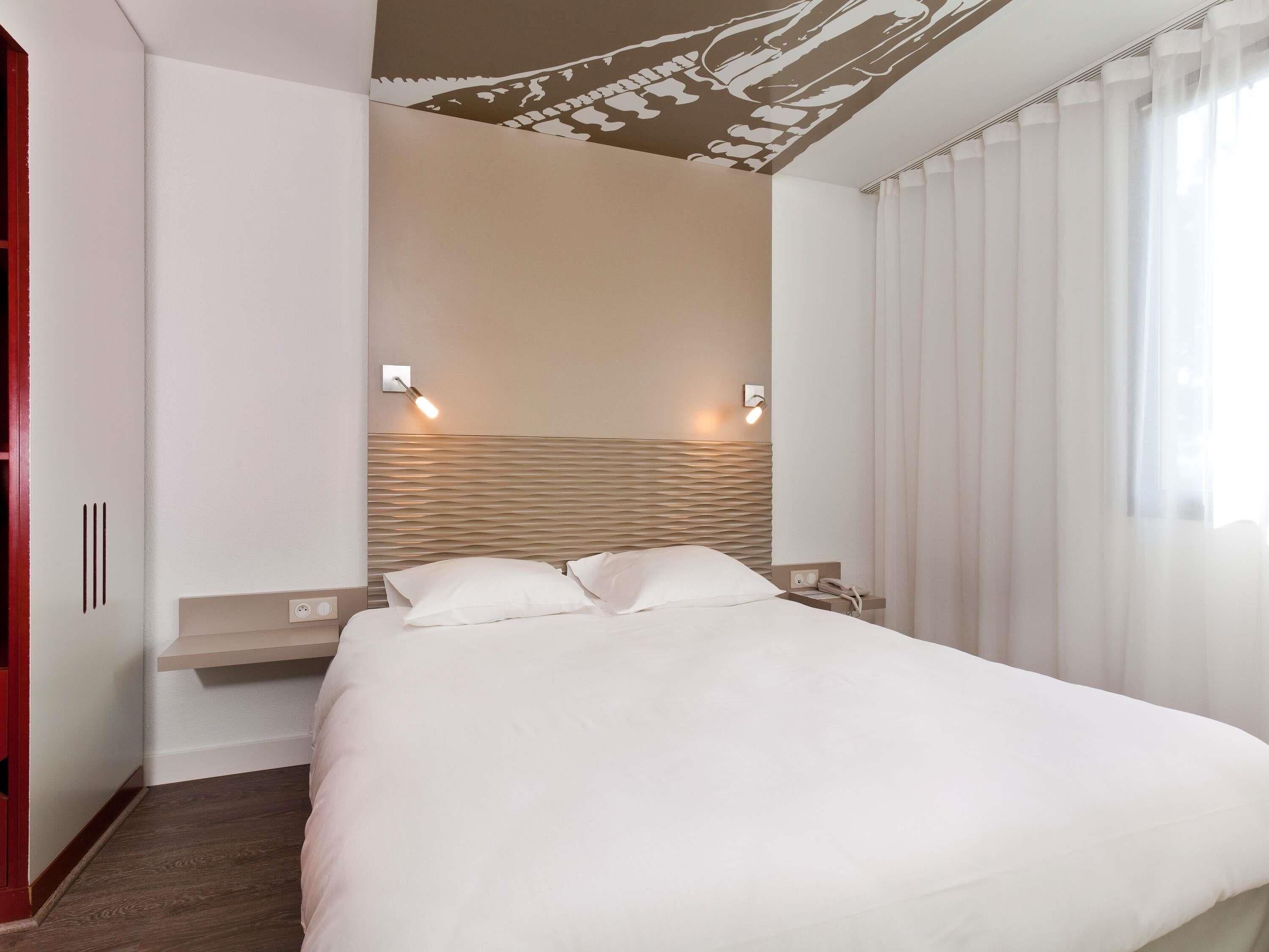 A modern hotel room with a large bed, two bedside tables, a headboard with a textured design, and a window with sheer curtains.