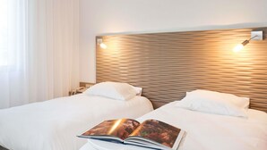 Une chambre d’hôtel avec deux lits, une tête de lit en bois, des lampes de chevet et une fenêtre avec des rideaux légers.