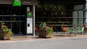 L’entrée d’un hôtel, avec une enseigne indiquant « ibis Styles Hotels » et la mention « Lille Aéroport » à proximité. On y trouve des espaces extérieurs aménagés avec des chaises et des tables.