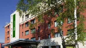 Un bâtiment moderne avec une enseigne verticale verte, de nombreuses fenêtres et un espace d’entrée abrité doté de sièges extérieurs.